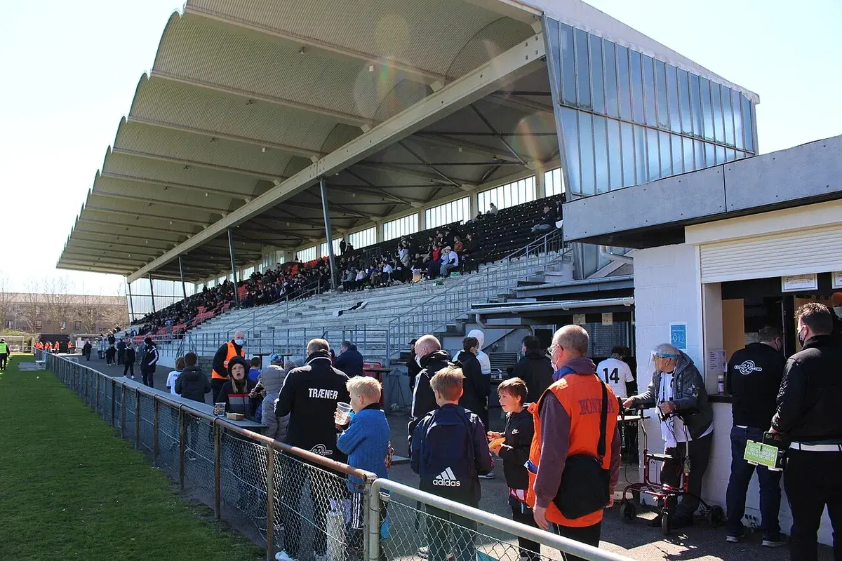 Exterior view of a sports stadium grandstand with spectators and people standing along the sideline in Copenhagen.