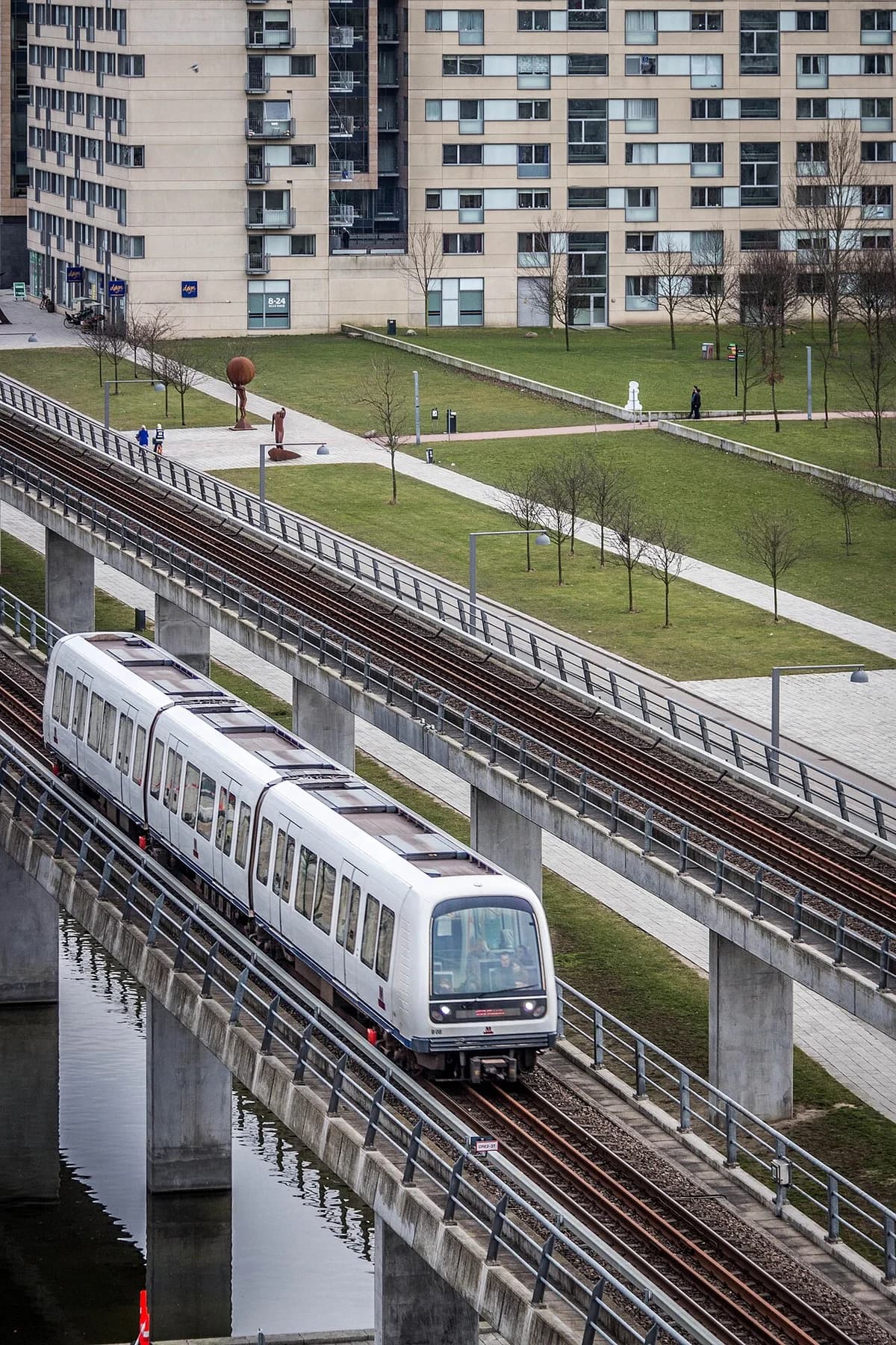 Copenhagen metro train on elevated track above water with urban buildings and park surroundings