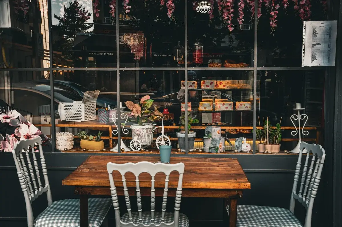 A cozy café table and chairs outside a decorated window display with plants and pastries.