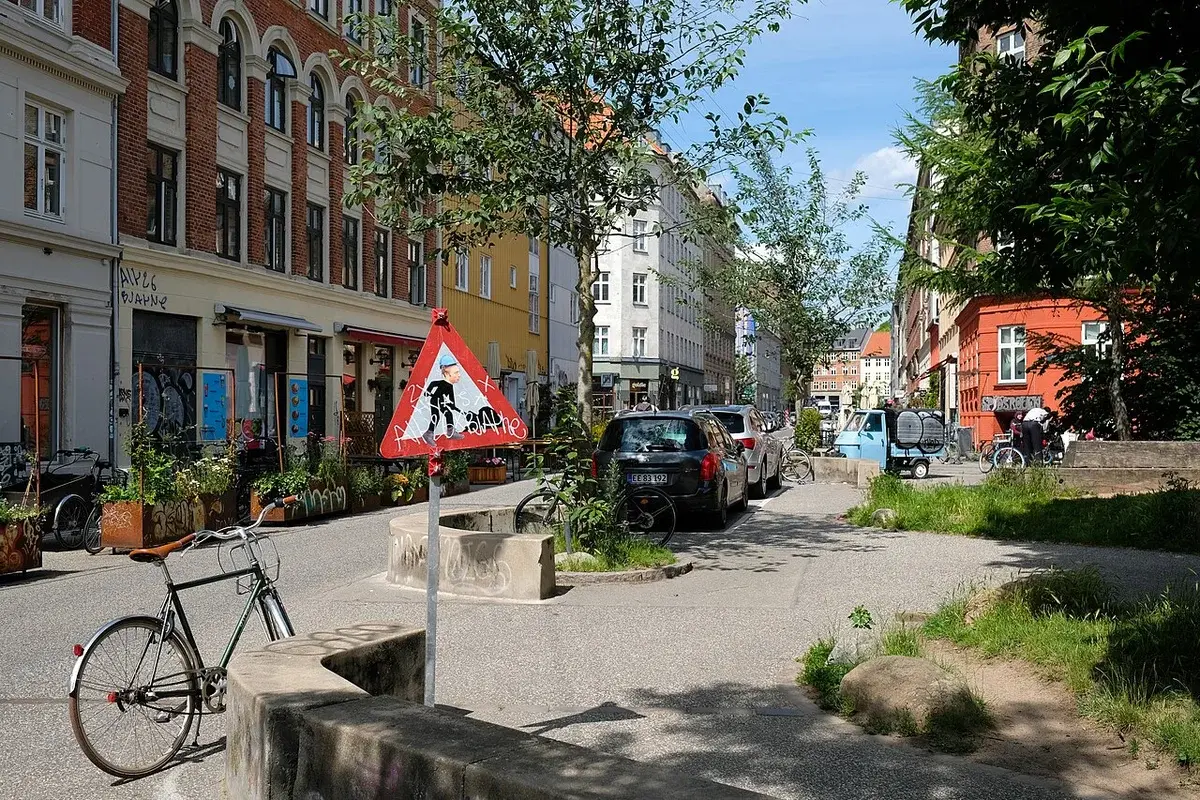 A sunny street scene in Frederiksberg with classic buildings, parked cars, bicycles, and trees.
