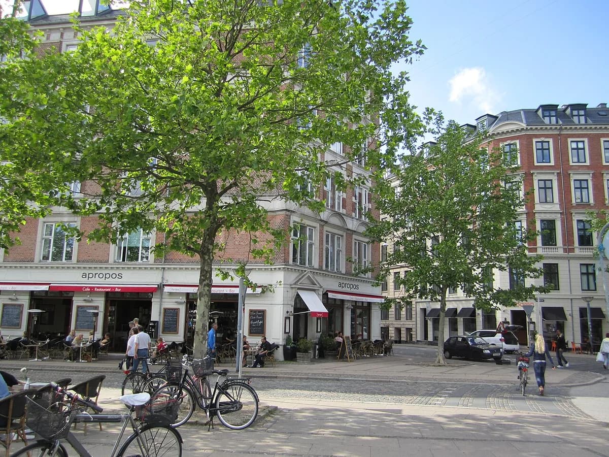 Sunny street scene in Vesterbro, Copenhagen with cafes, apartment buildings, trees, bicycles, and pedestrians.