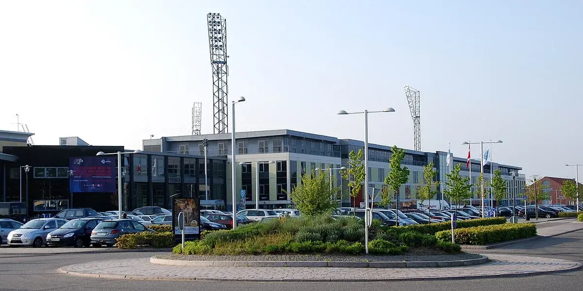 Exterior view of Viborg Stadium with parked cars, Danish flags, and tall floodlight towers.