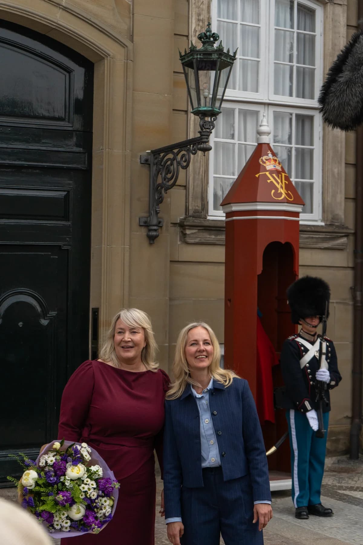 Two women stand together outside a government building, one holding a bouquet, with a ceremonial guard in the background.