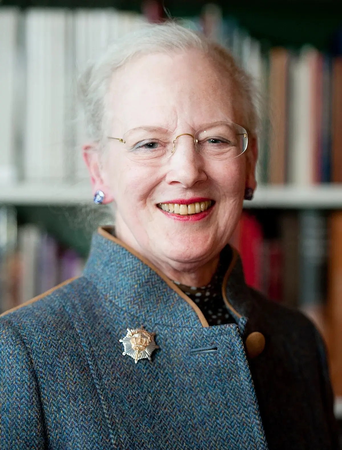 Close-up portrait of Queen Margrethe II smiling in front of bookshelves.