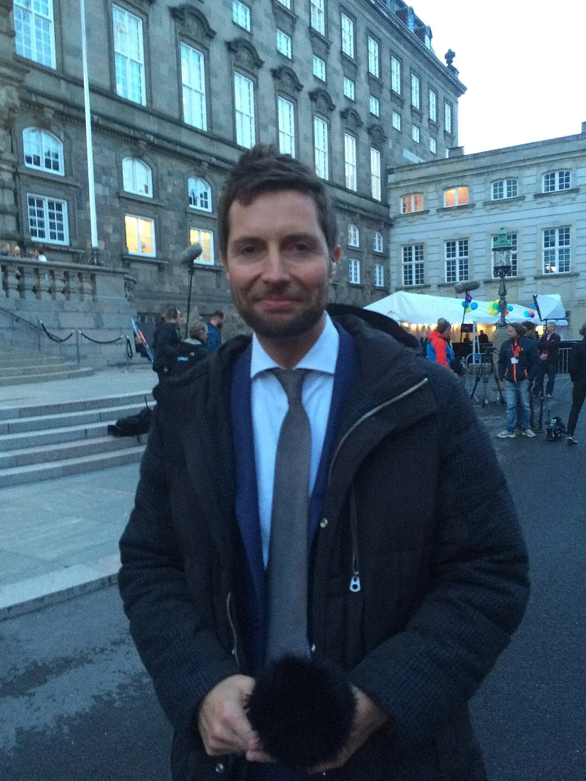 A man in a suit and jacket stands outside a historic building during election coverage.