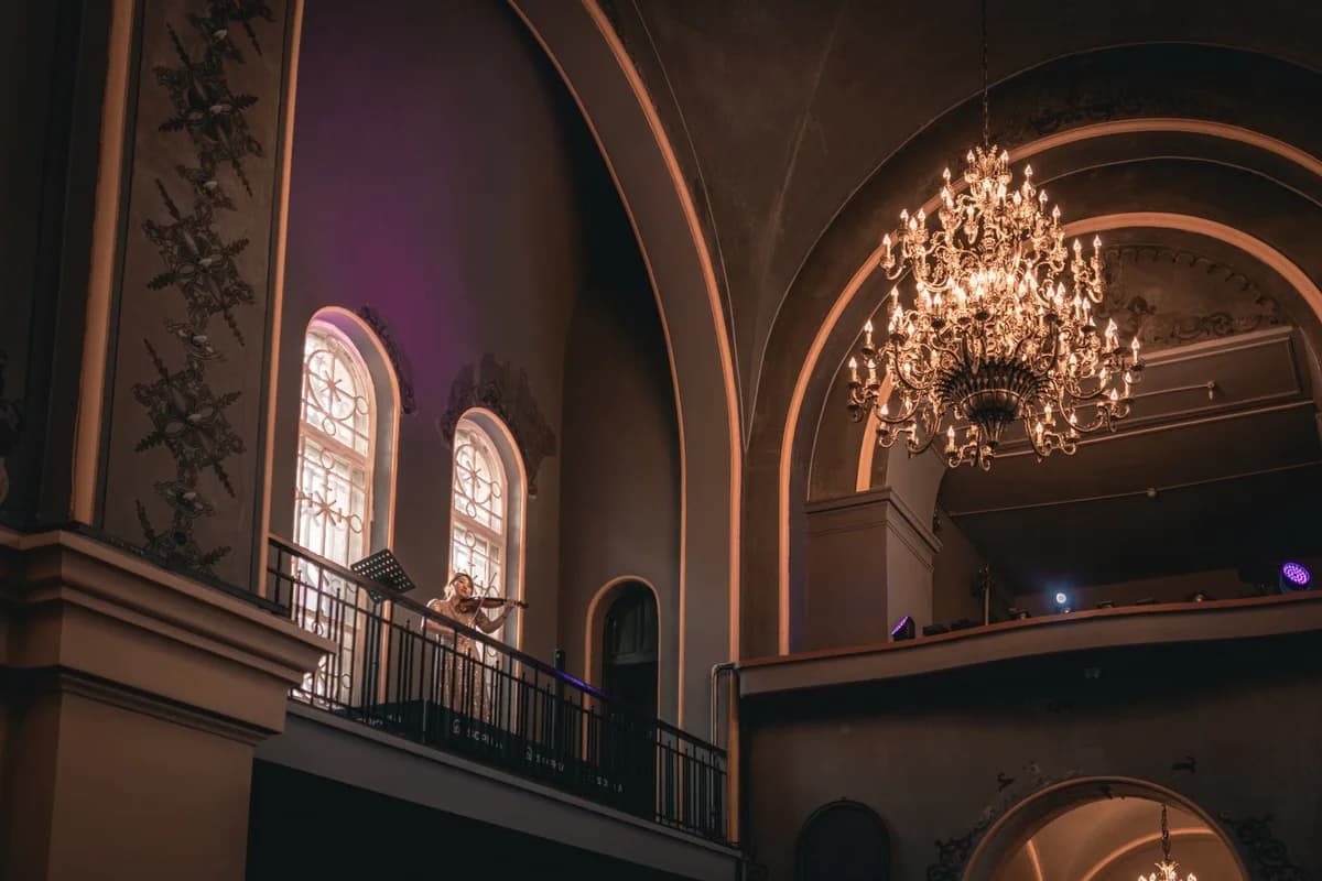 A violinist performs on a balcony in a grand hall beneath an ornate chandelier.
