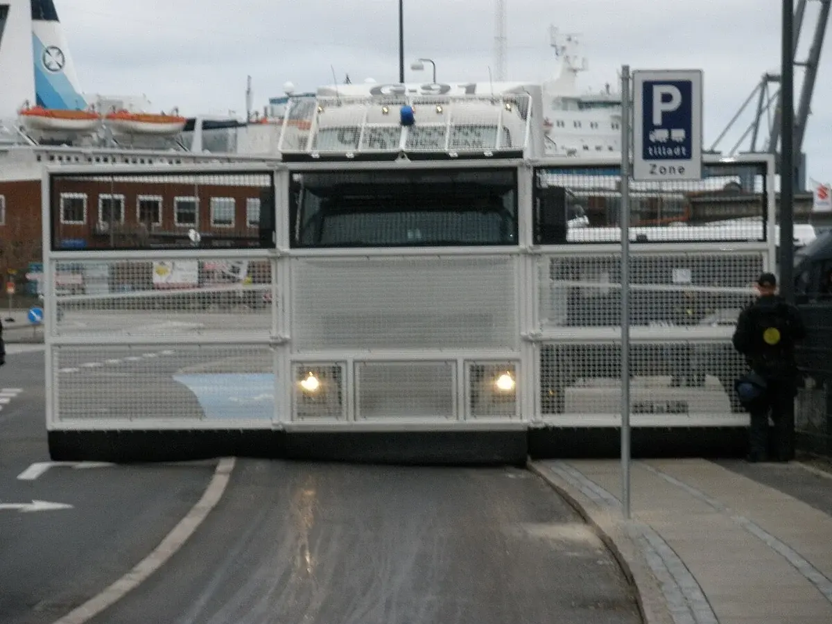 A white Volvo police truck with a reinforced barrier is shown on a Copenhagen street next to a parking sign, with a police officer standing beside it.