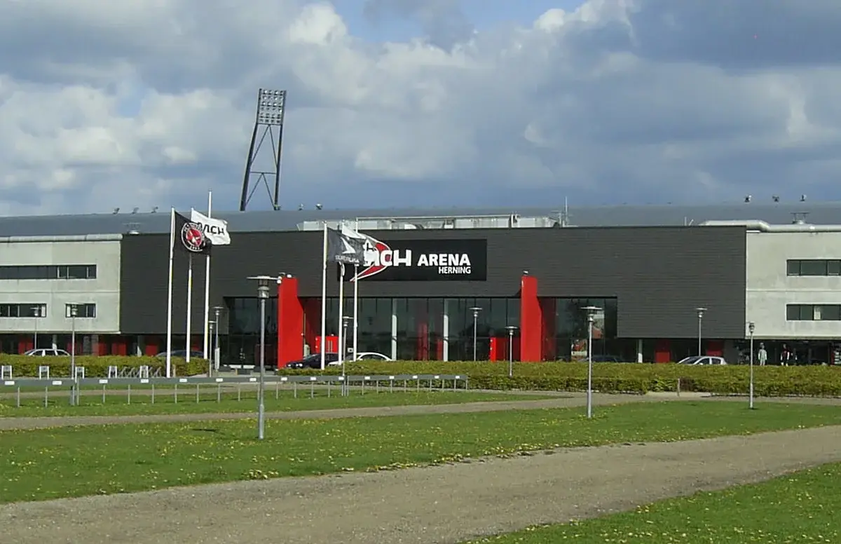 Exterior of MCH Arena in Herning with flags and a stadium light tower under a cloudy sky.