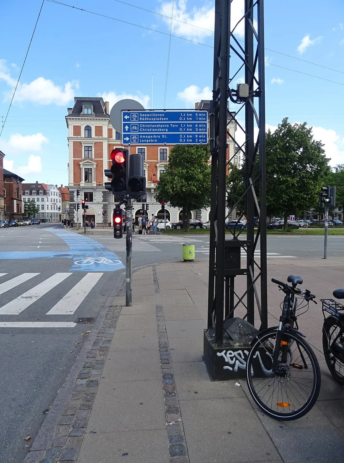 A Copenhagen street corner with a bicycle direction sign, traffic lights, and parked bikes.