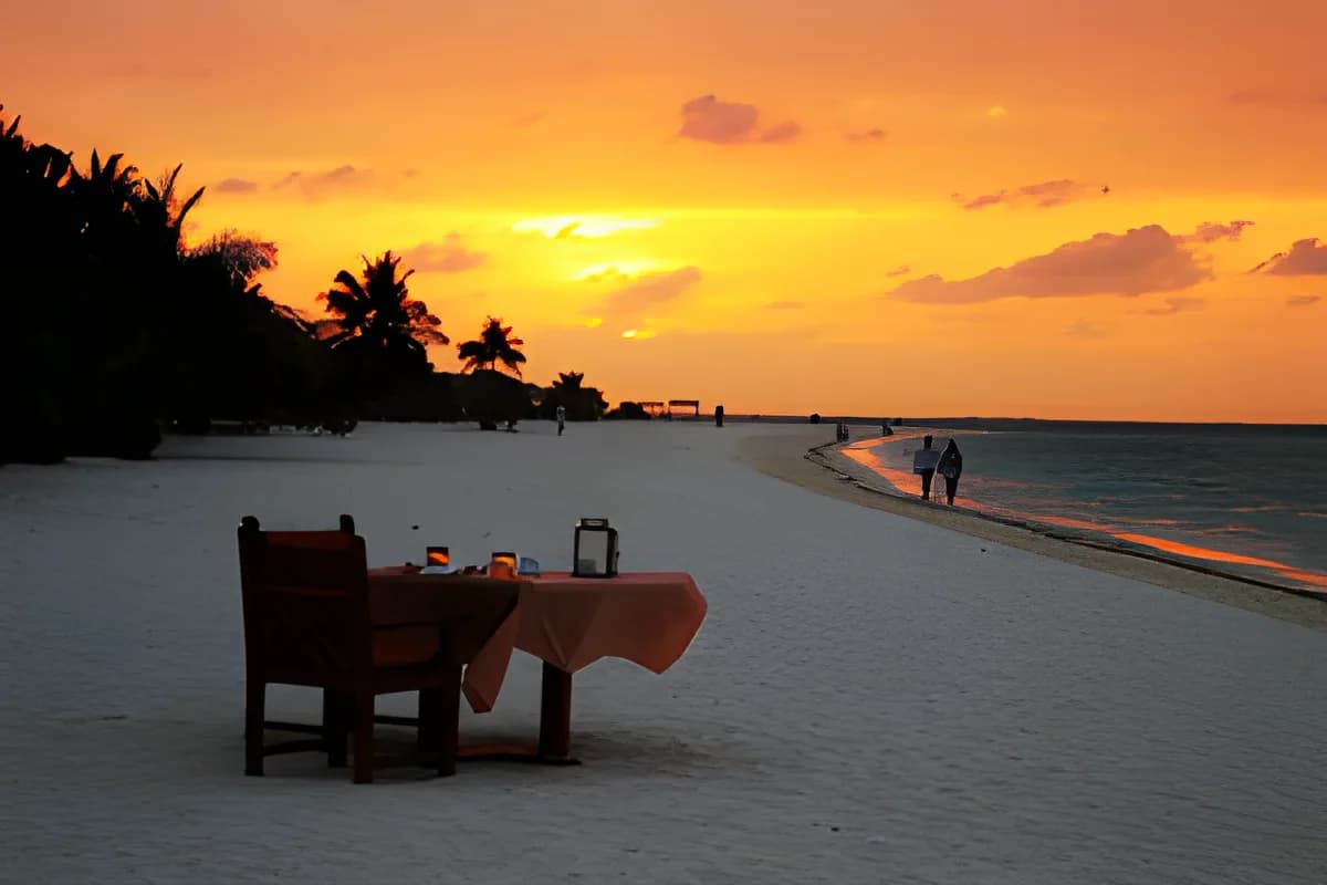 Sunset over a Maldivian beach with palm trees, a resort dining table, and people walking by the water.