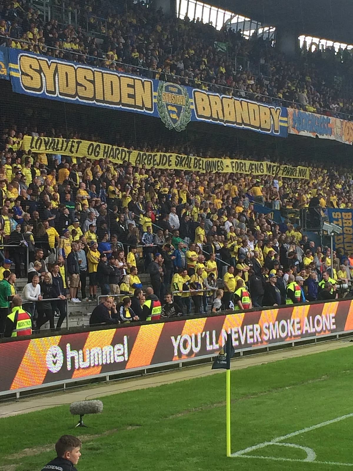 Crowded Brøndby IF supporters in the South Stand at a football match, with yellow banners and pitchside advertising visible.