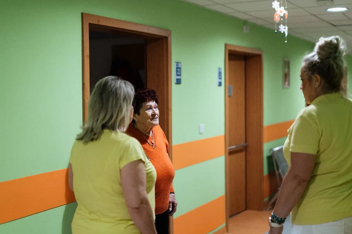 An older woman speaks with two female care workers in a green-and-orange hallway.