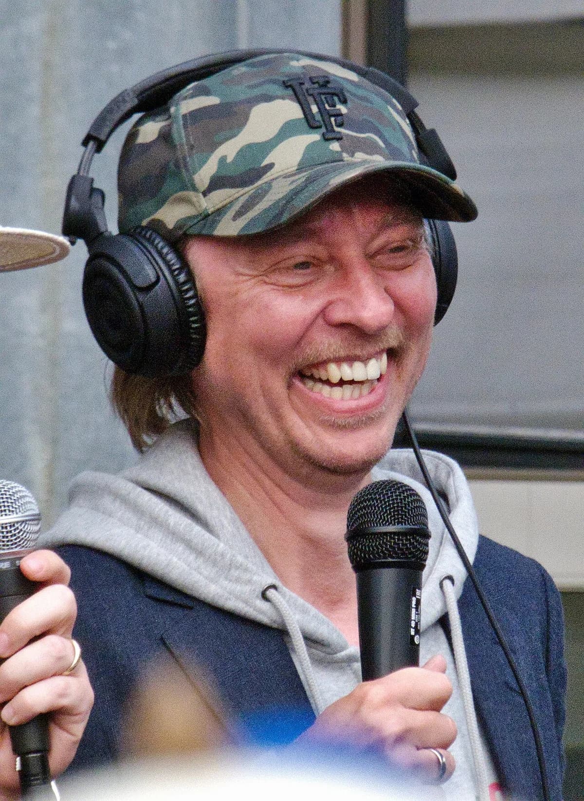 A smiling man wearing headphones and a camouflage cap speaks into a microphone.