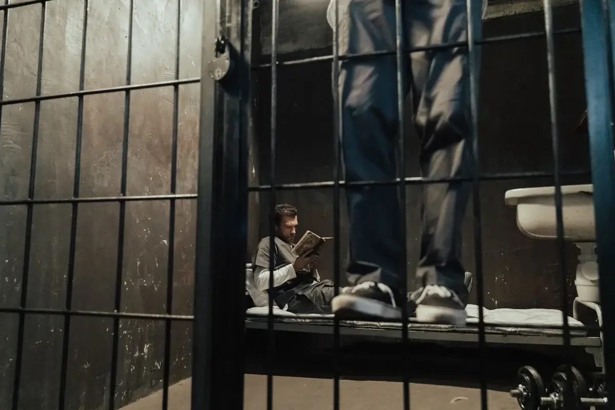 A man reads a book in a jail cell while another person stands nearby behind bars.