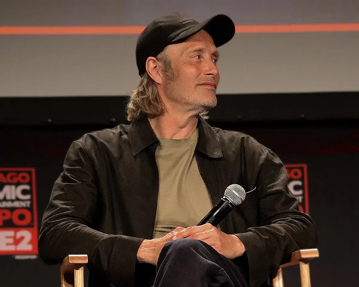 Mads Mikkelsen seated onstage holding a microphone at a C2E2 panel.