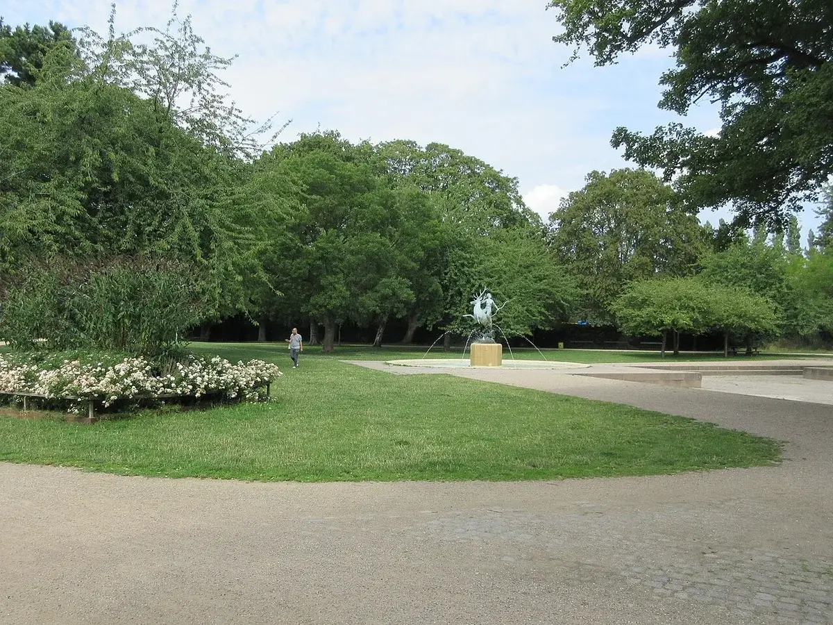 A park-like square with trees, grass, flower beds, and a fountain sculpture.